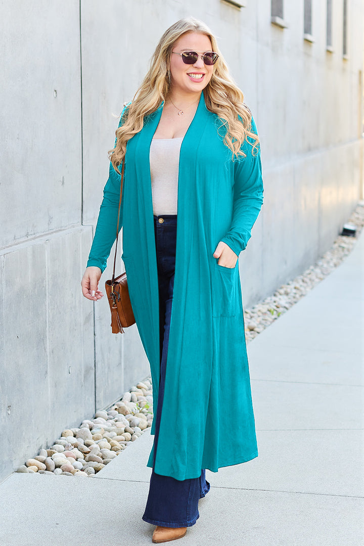 A woman with long blonde hair wearing sunglasses, a Basic Bae Full Size Open Front Long Sleeve Cover Up in teal, a white shirt, dark blue jeans, and a brown bag walks down a sidewalk next to a modern building. She is smiling and looking relaxed.