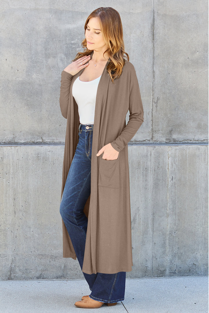 A woman stands against a concrete wall, wearing the Basic Bae Full Size Open Front Long Sleeve Cover Up in brown, paired with a white top and blue jeans. She rests her hand on her neck, looking to the side pensively.