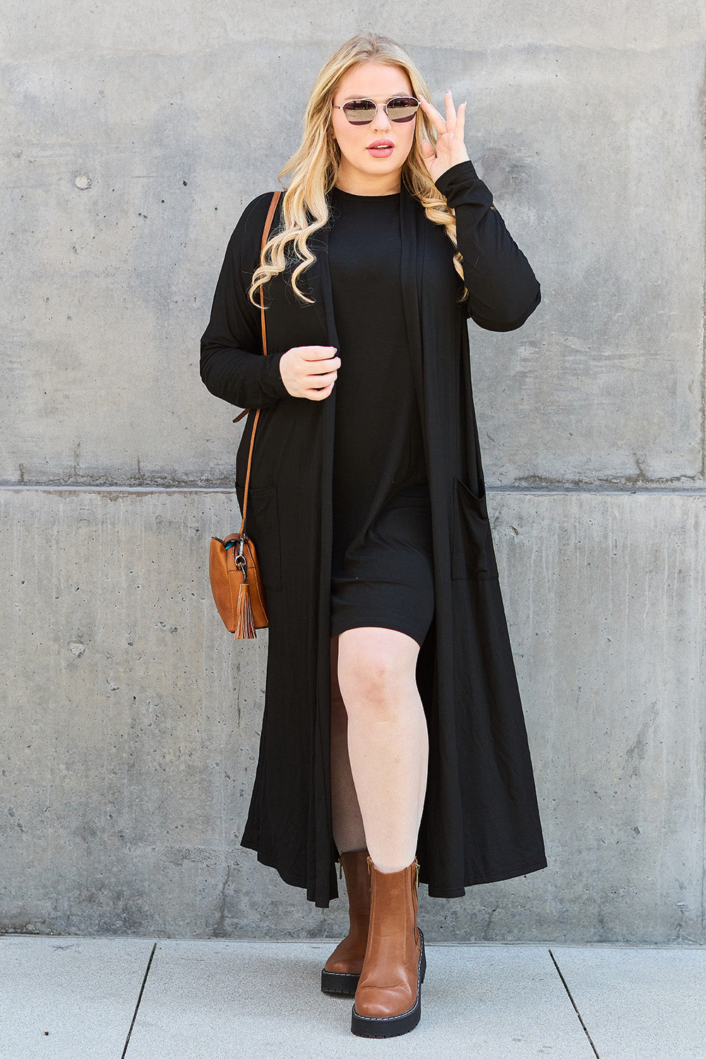 A person with long blonde hair wears a black dress made from stretchy material, paired with the Basic Bae Full Size Open Front Long Sleeve Cover Up in black, and brown ankle boots. They carry a brown purse and wear sunglasses, posing confidently against a concrete wall.