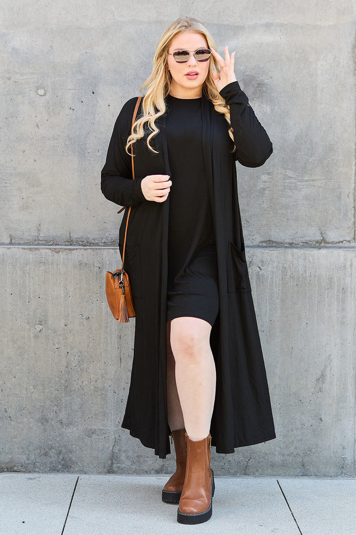 A person with long blonde hair wears a black dress made from stretchy material, paired with the Basic Bae Full Size Open Front Long Sleeve Cover Up in black, and brown ankle boots. They carry a brown purse and wear sunglasses, posing confidently against a concrete wall.