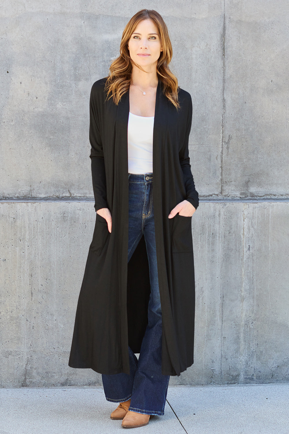 A person wearing the Basic Bae Full Size Open Front Long Sleeve Cover Up in black over a white top and blue jeans stands against a concrete wall. With hands tucked into the pockets of the sheer design, they complete the look with brown shoes.