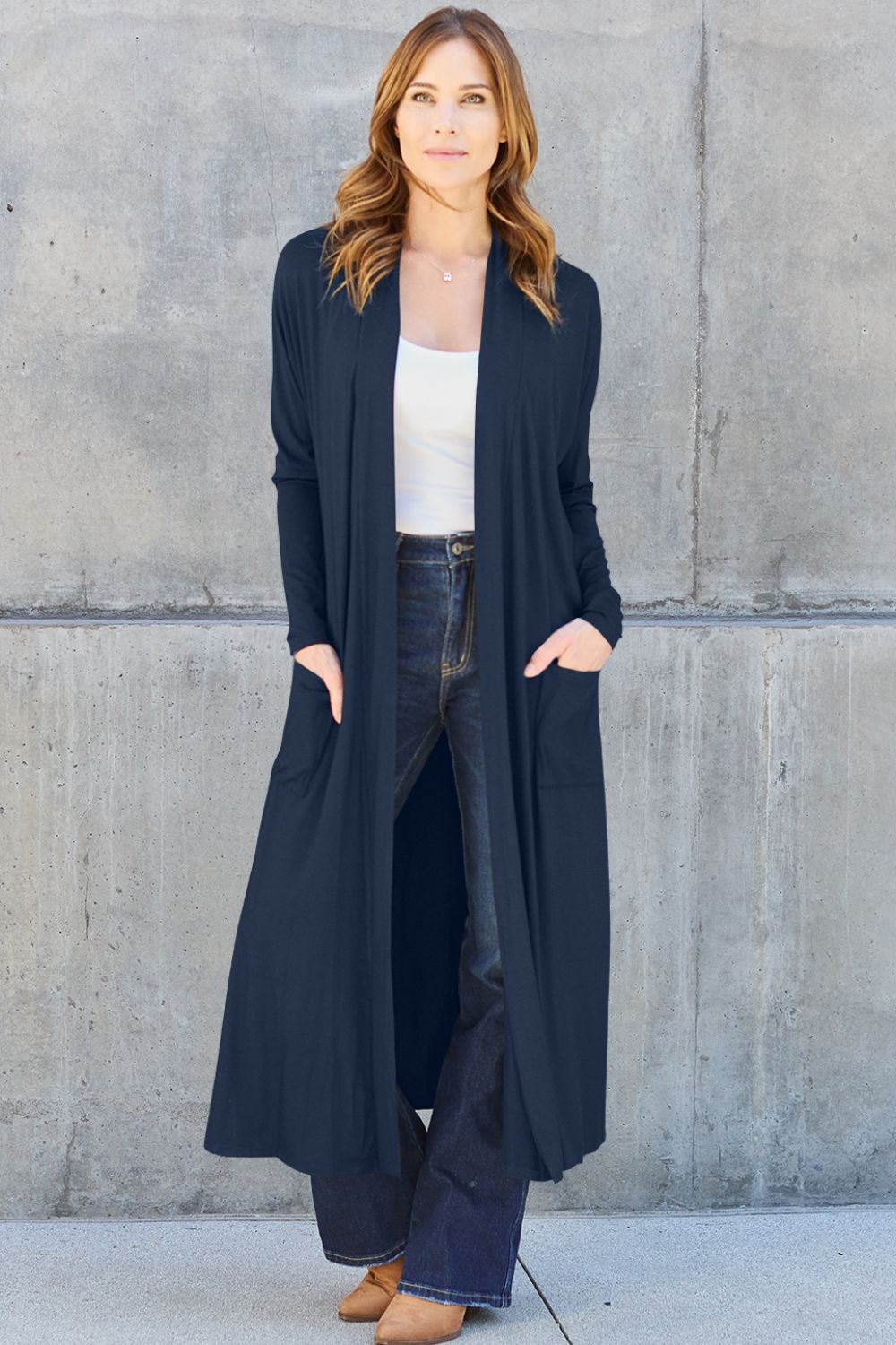A woman stands against a concrete wall wearing the Basic Bae Full Size Open Front Long Sleeve Cover Up in navy, paired with a white top, dark jeans, and brown shoes. Her hands rest in the pockets of the stretchy material, complementing her long, light brown hair. The overall look is casual and stylish.