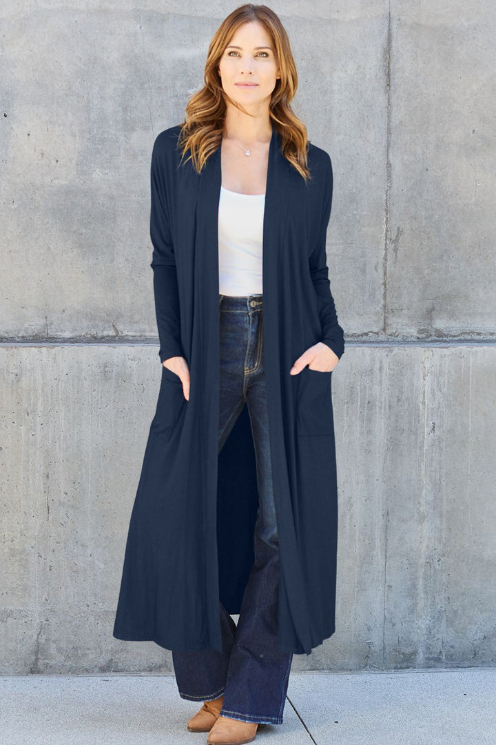 A woman stands against a concrete wall wearing the Basic Bae Full Size Open Front Long Sleeve Cover Up in navy, paired with a white top, dark jeans, and brown shoes. Her hands rest in the pockets of the stretchy material, complementing her long, light brown hair. The overall look is casual and stylish.