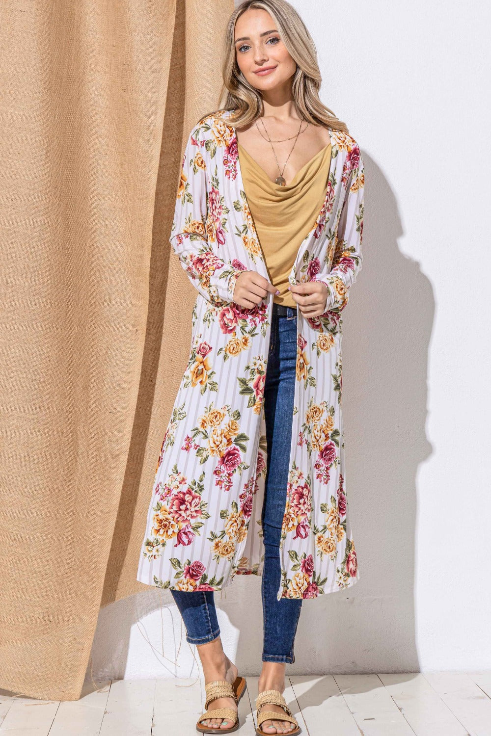 A person stands against a light background, wearing the And The Why Floral Kimono Open Front Longline Cardigan over a mustard blouse and blue jeans. They are also wearing sandals and have long, light-colored hair. A beige curtain is partially visible beside them.