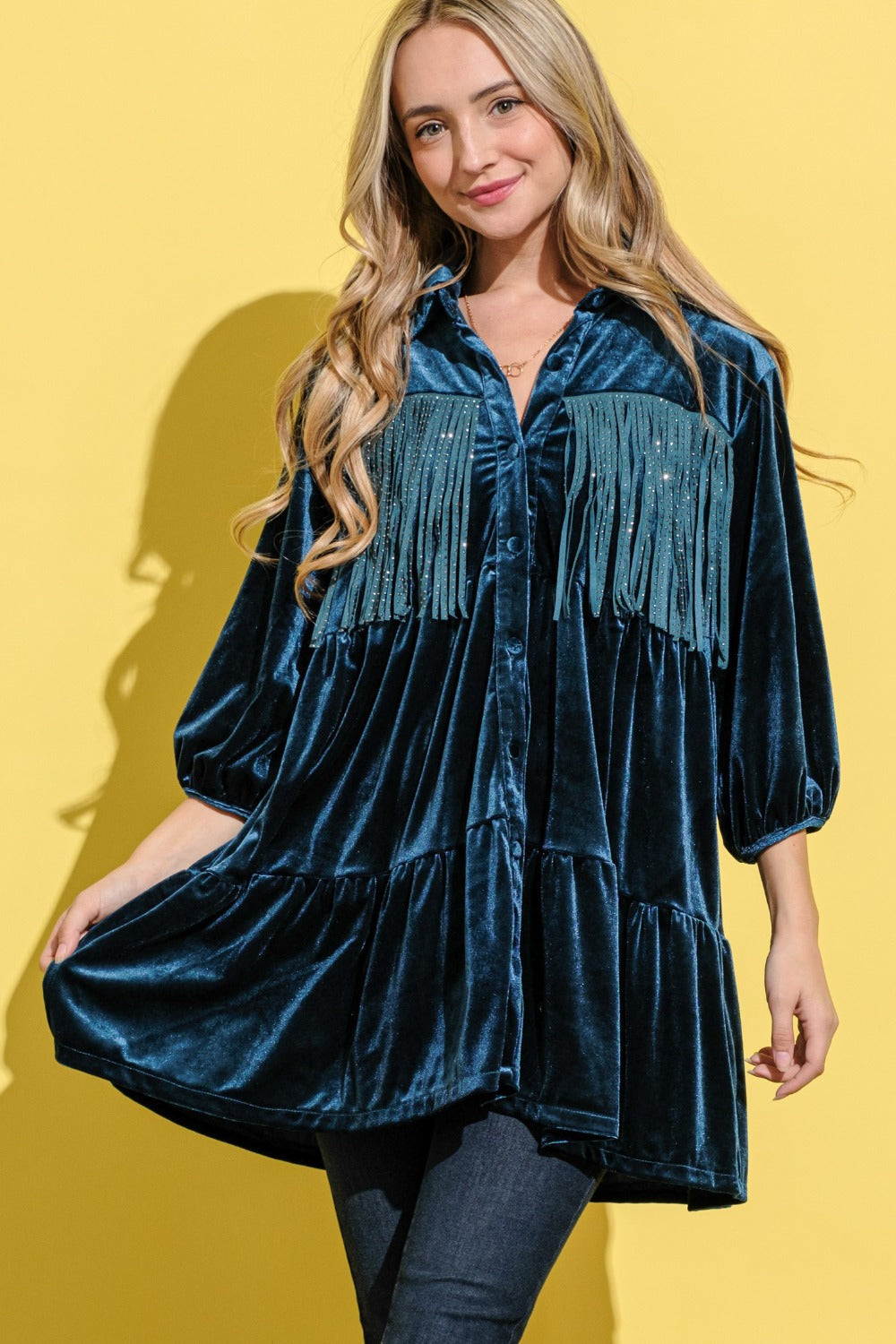 Against a yellow backdrop, a woman smiles at the camera in The Why Fringe Detailed Velvet Shirt Dress. This dark blue dress showcases three-quarter sleeves, fringe detailing, and a tiered design. Her long blonde hair flows in loose waves.