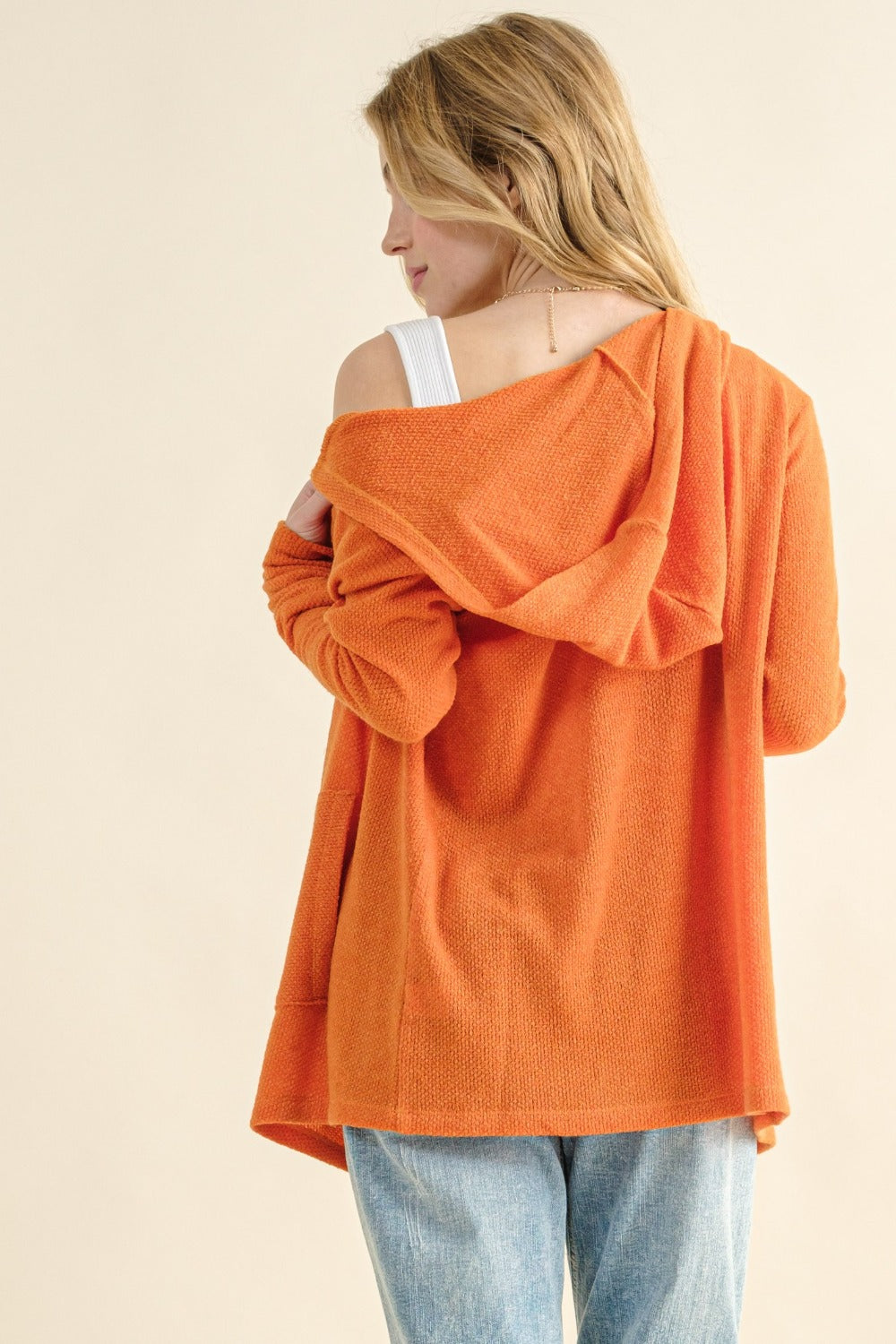 A woman wearing the "And The Why Full Size Thermal Hooded Open Front Cardigan with Pockets" stands with her back to the camera, looking left. The cozy layering sweater drapes off one shoulder, showing a white strap underneath. She has long blonde hair and is paired with light blue jeans against a neutral background.