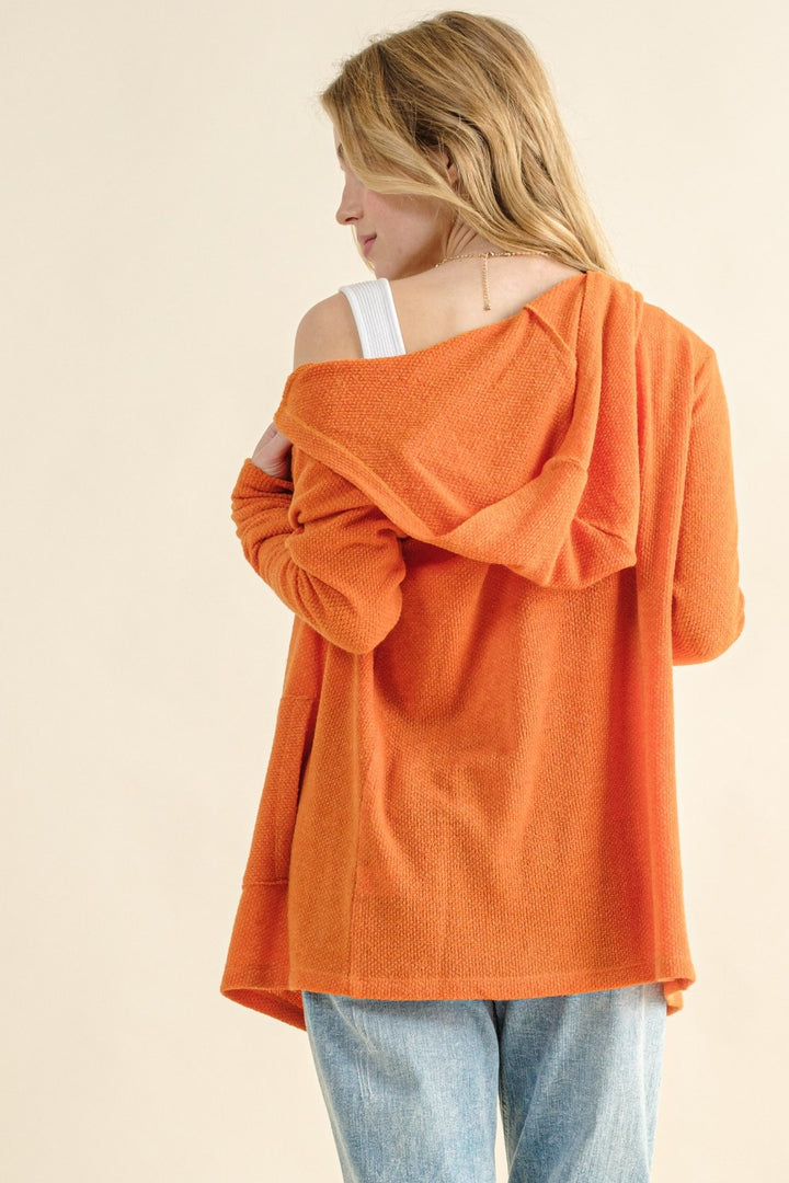 A woman wearing the "And The Why Full Size Thermal Hooded Open Front Cardigan with Pockets" stands with her back to the camera, looking left. The cozy layering sweater drapes off one shoulder, showing a white strap underneath. She has long blonde hair and is paired with light blue jeans against a neutral background.