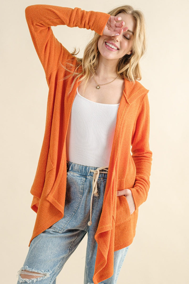 A person stands smiling, wearing the And The Why Full Size Thermal Hooded Open Front Cardigan with Pockets in a warm orange color over a white tank top and blue jeans. One hand is casually in their pocket, while the other arm is raised, gently touching their forehead against a neutral beige background.