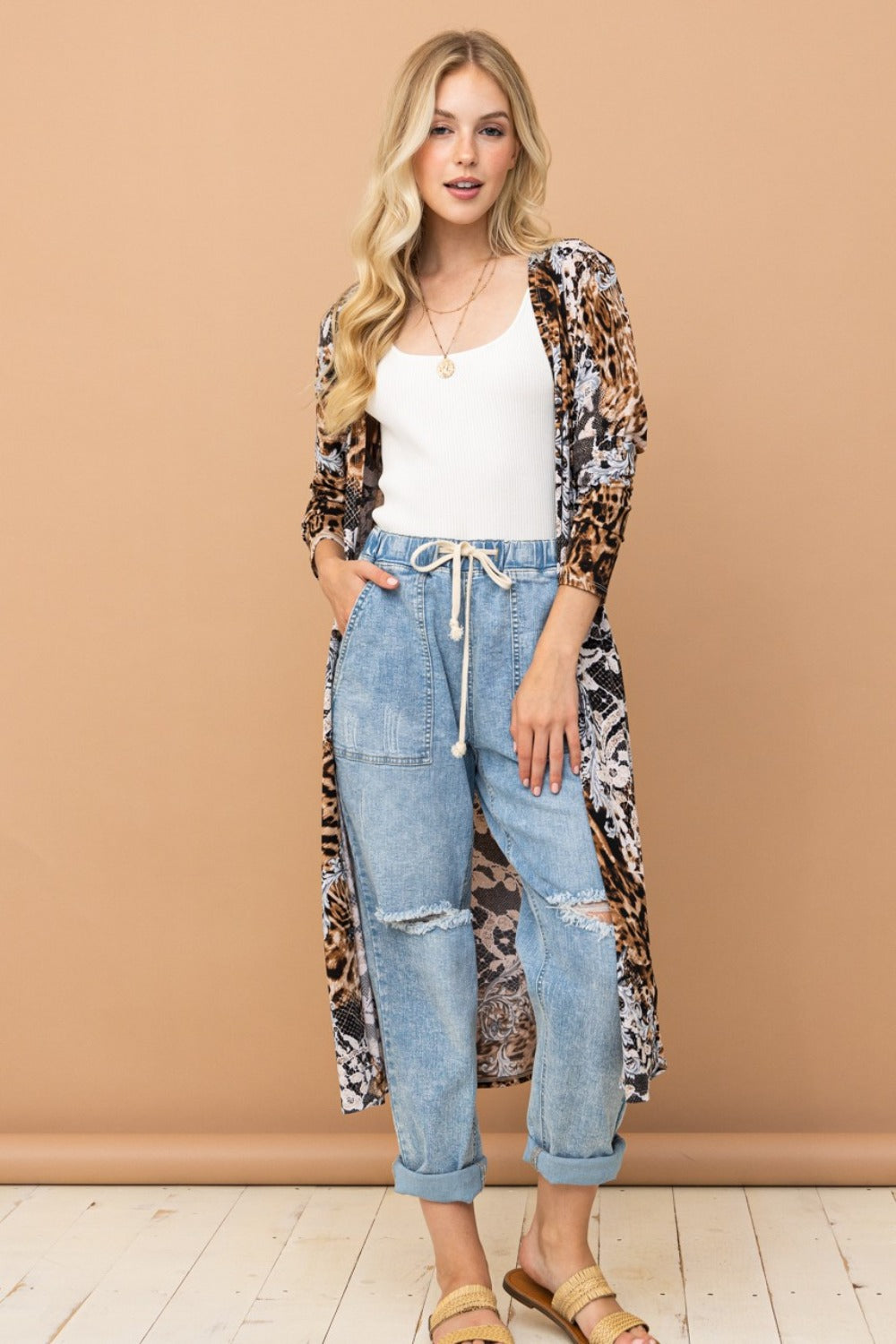 A woman in a white tank top and light blue jeans with a drawstring stands against a beige background. She is wearing the And The Why Leopard Kimono Open Front Longline Cardigan paired with gold sandals. Her long, wavy hair cascades down as she smiles slightly.
