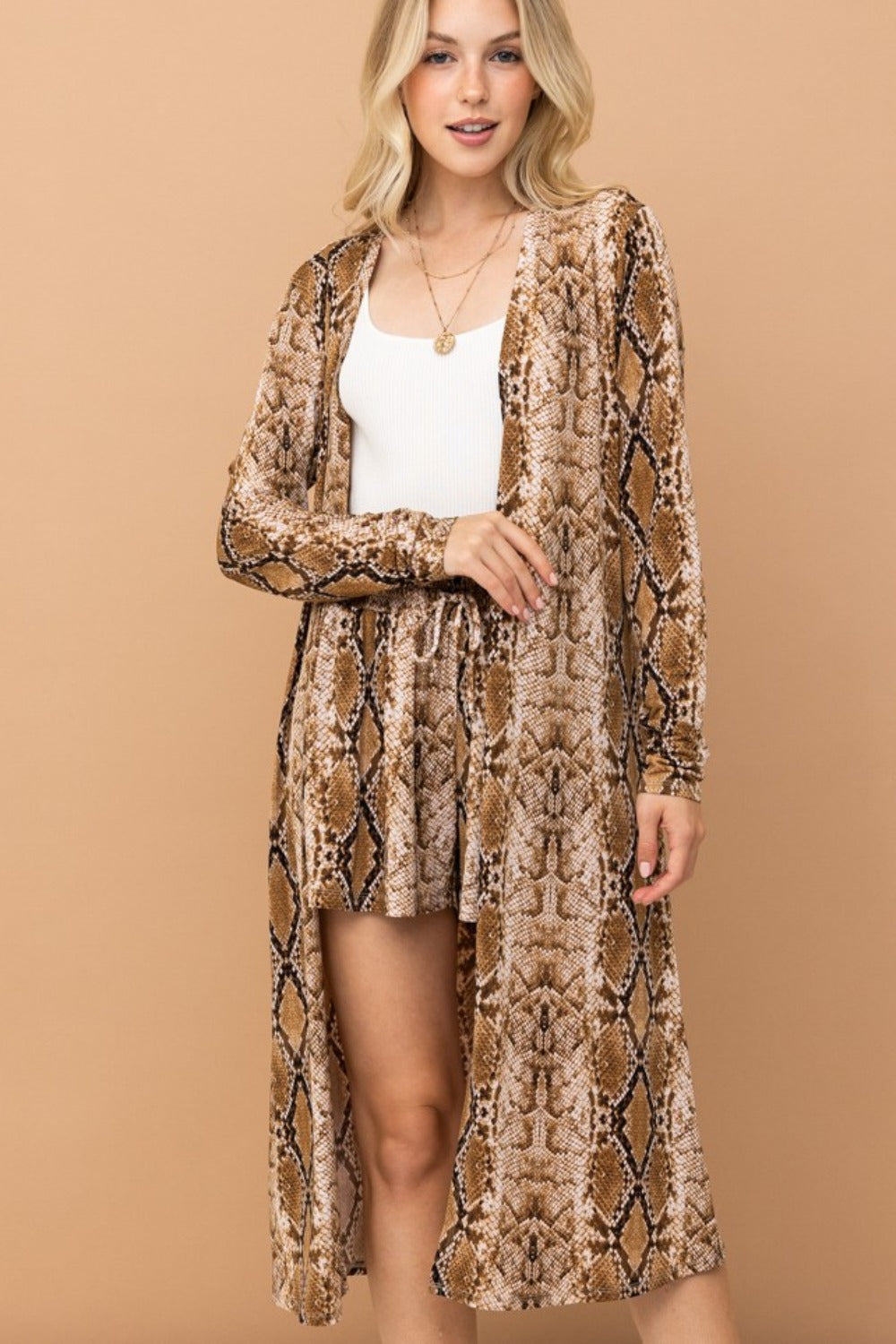 A woman poses against a beige background, wearing the And The Why Snake Print Kimono Open Front Longline Cardigan over a white top and shorts. She has long blonde hair and wears minimal jewelry, including a necklace. Her expression is relaxed and confident.