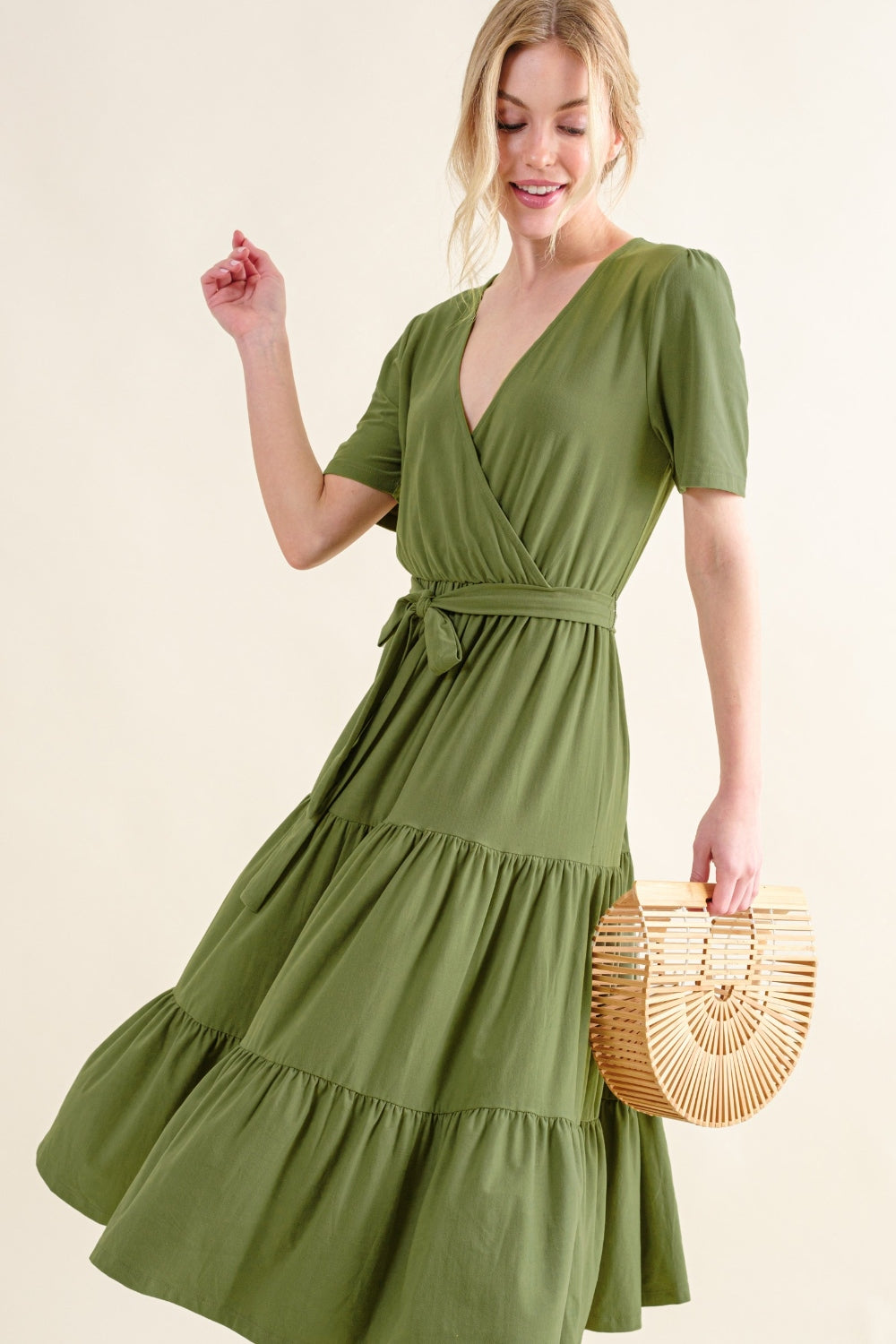 A person dressed in the And The Why Soft Short Sleeve Tiered Midi Dress, featuring a flowy silhouette and a green hue, is holding a round wooden handbag. They are smiling with their hair tied back against a plain, light background.