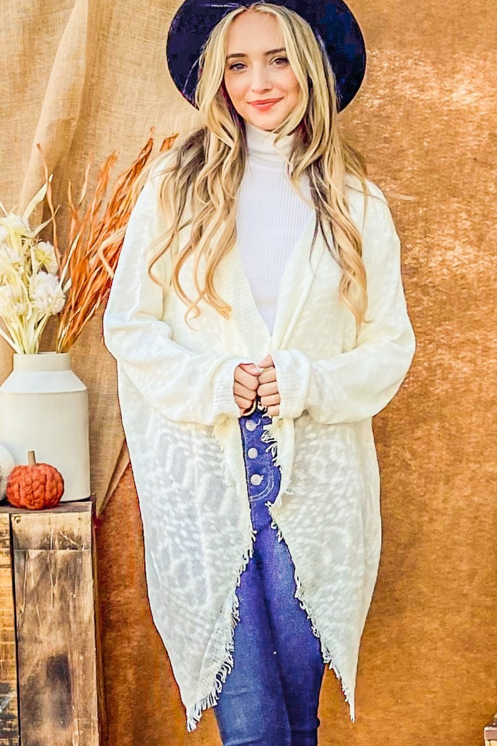 Dressed in an And The Why Textured Knit Fringe Hem Cardigan over a turtleneck and jeans, a person with long blonde hair and a black hat embodies bohemian flair. They are posed in front of a beige backdrop, where a small table holds a vase filled with flowers and a pumpkin.
