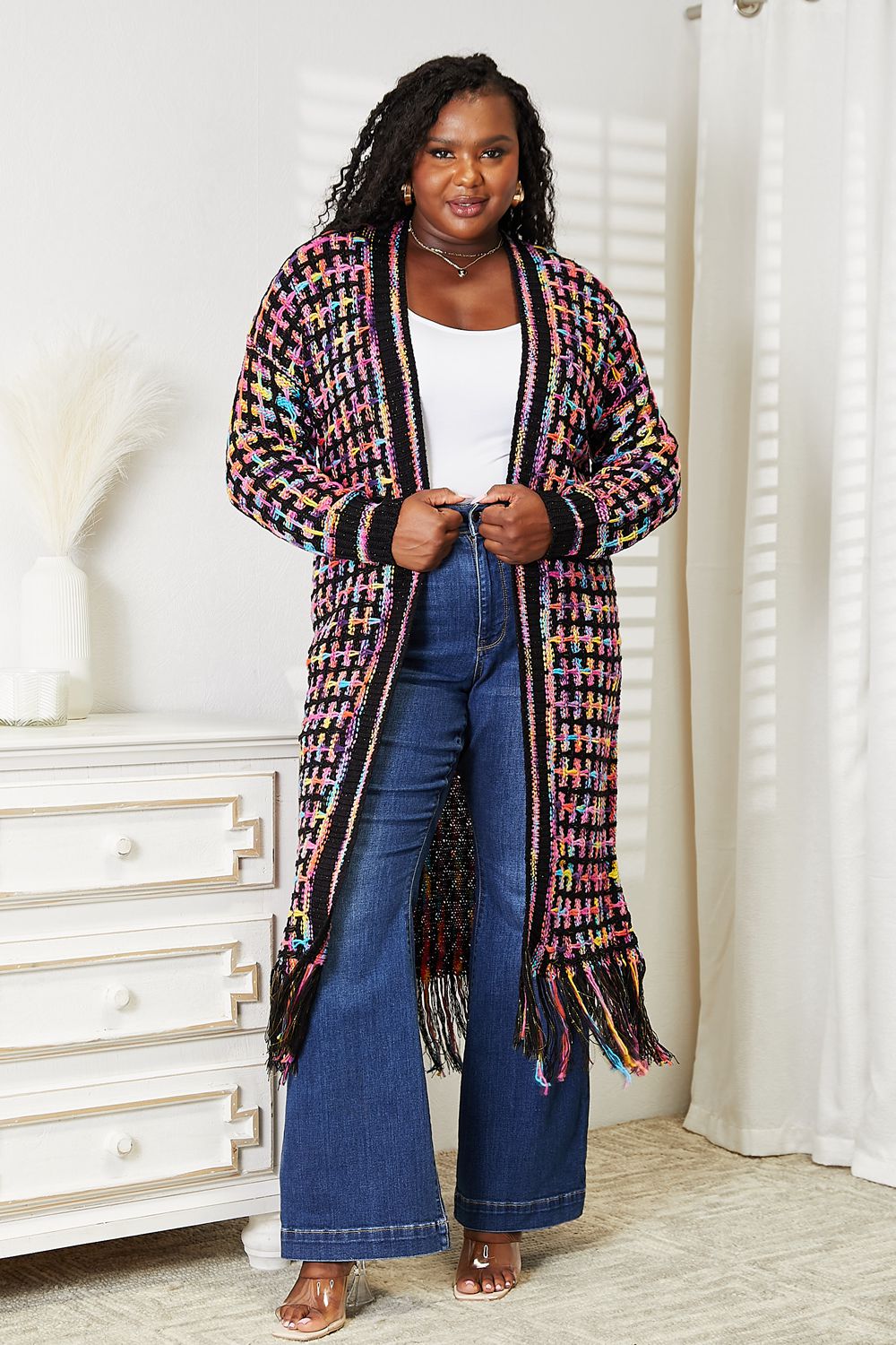 A person is wearing the Angel Wings Full Size Multicolored Open Front Fringe Hem Cardigan over a white top and blue wide-leg jeans. They are standing in a room with a white dresser and vase in the background, smiling with hands in pockets, exuding effortless bohemian style.
