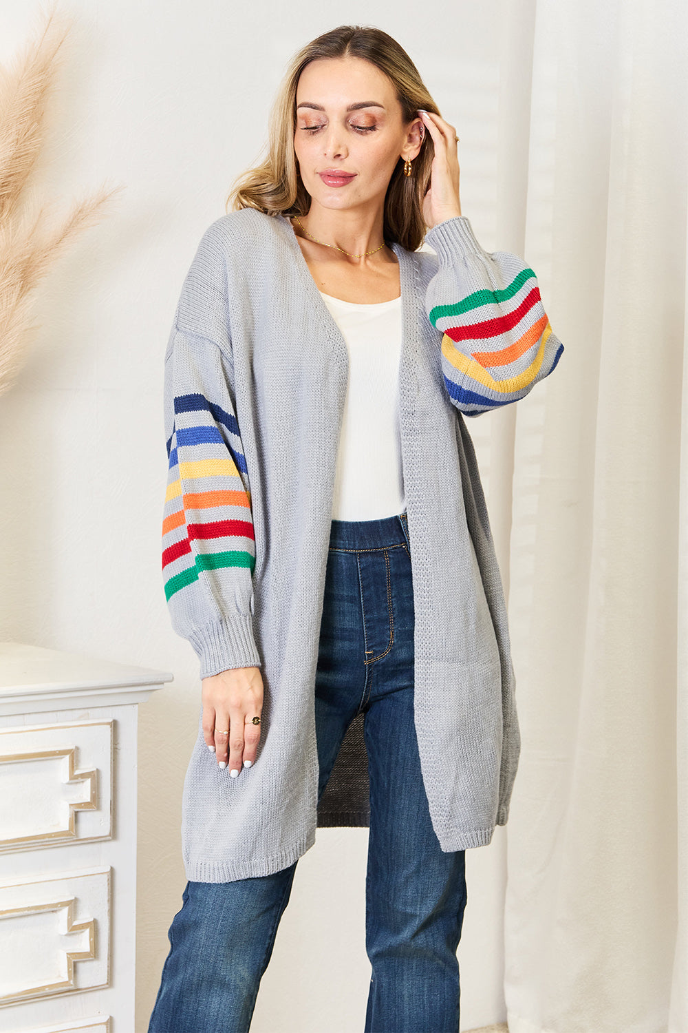 Indoors, an individual dons the stylish Angel Wings Multicolored Stripe Open Front Longline Cardigan over a white top and blue jeans. With long hair, they gaze slightly to the side. In the backdrop, a white cabinet is accompanied by decorative grasses.