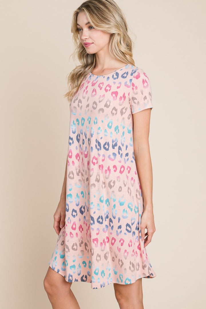 A woman wearing a BOMBOM Leopard Round Neck Short Sleeve Dress, featuring a colorful leopard print in pink, blue, and beige tones, stands against a soft, neutral background.