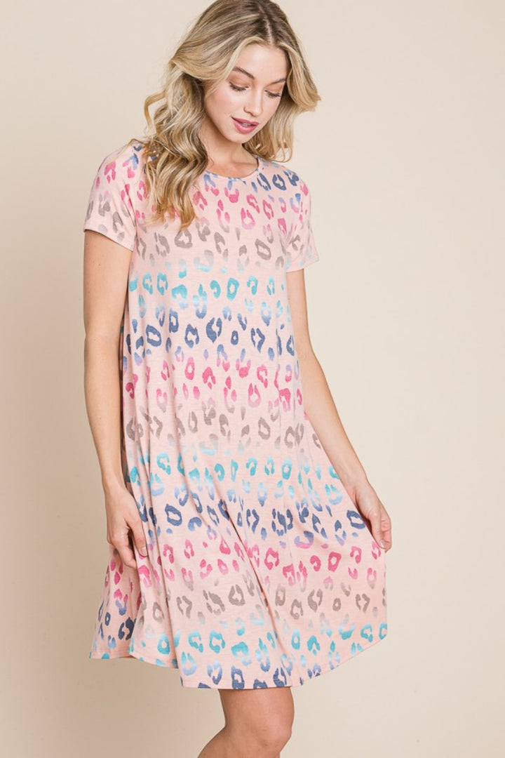 A woman elegantly models the BOMBOM Leopard Round Neck Short Sleeve Dress, showcasing its light pink base with a vibrant multicolored leopard print in blue, pink, and tan against a plain beige backdrop.