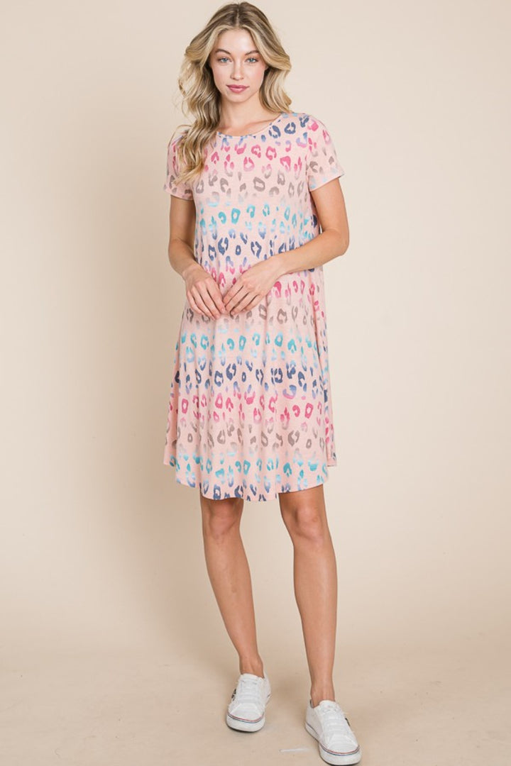 A person stands against a beige background wearing the BOMBOM Leopard Round Neck Short Sleeve Dress, knee-length with short sleeves. They pair it with white sneakers, and their long, wavy hair cascades down, enhancing the chic ensemble.
