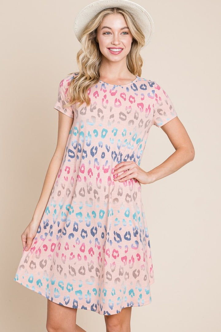 A woman in a vibrant BOMBOM Leopard Round Neck Short Sleeve Dress smiles while wearing a straw hat. Her hair cascades freely against a plain, light beige background.