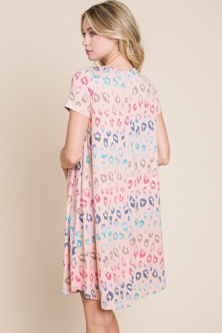 A woman models the BOMBOM Leopard Round Neck Short Sleeve Dress, showcasing its loose fit and vibrant pink, blue, and beige leopard print. She stands sideways against a plain background, looking over her shoulder.