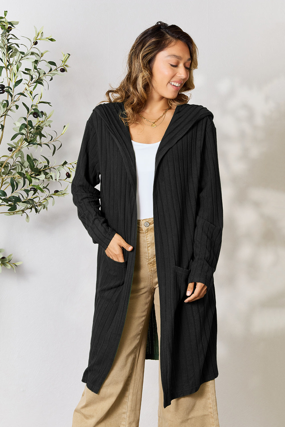 A person stands smiling, wearing the Basic Bae Full Size Hooded Sweater Cardigan in black over a white top and beige pants. They have long, wavy hair and hold the cardigan's pocket with one hand. The cardigan beautifully complements the green leafy plant in the background against a plain wall.