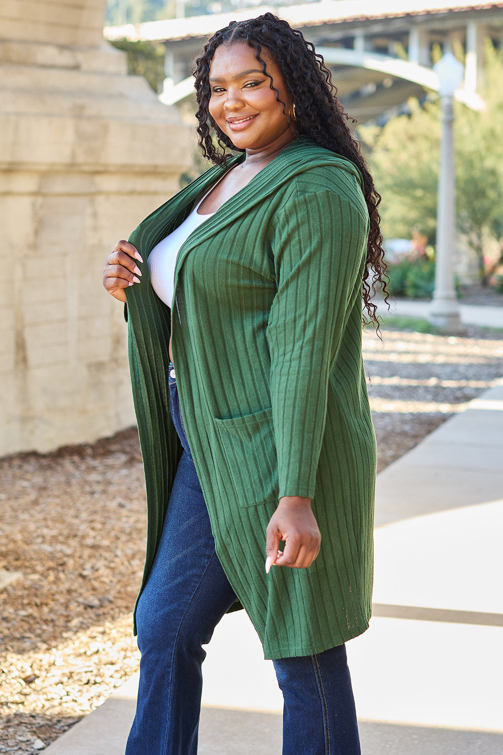 A person smiling and standing outdoors, wearing the Basic Bae Full Size Hooded Sweater Cardigan in green over a white top and blue jeans. They have long, curly hair and are holding the cardigan with one hand. Stone structures and a pathway are visible in the background.