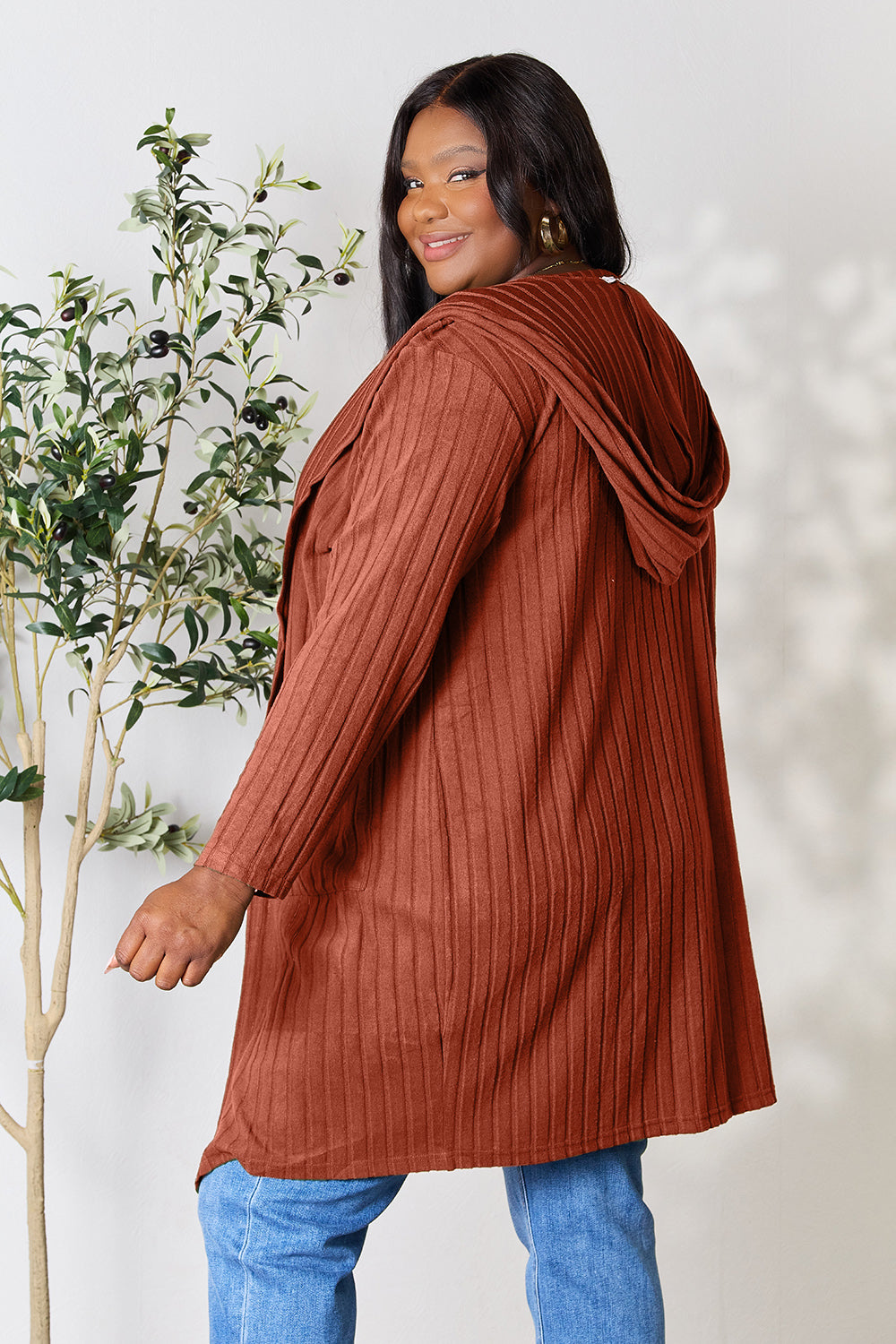 A person wearing the Basic Bae Full Size Hooded Sweater Cardigan in a ribbed, rust color and blue jeans smiles while standing next to a small leafy plant. This pocketed sheer cardigan is perfect for a casual day out. The background is a simple white wall.