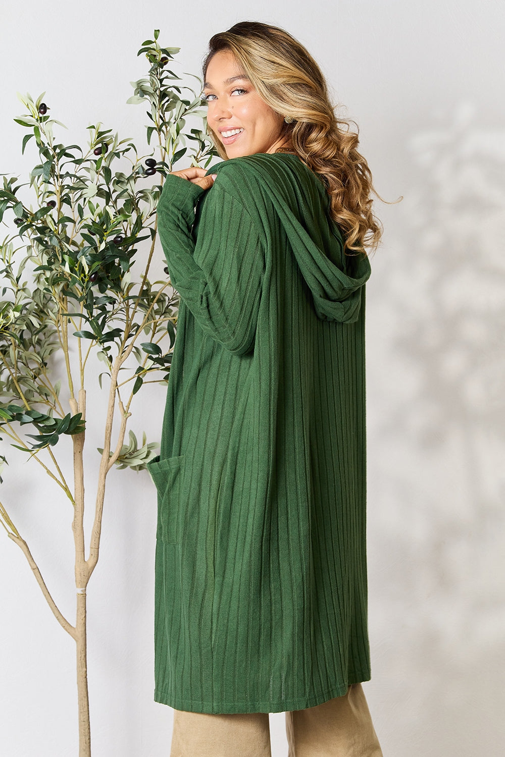 A person with long hair stands smiling, donned in the Basic Bae Full Size Hooded Sweater Cardigan in green. They are glancing over their shoulder next to a tall potted plant, set against a plain, light-colored wall.