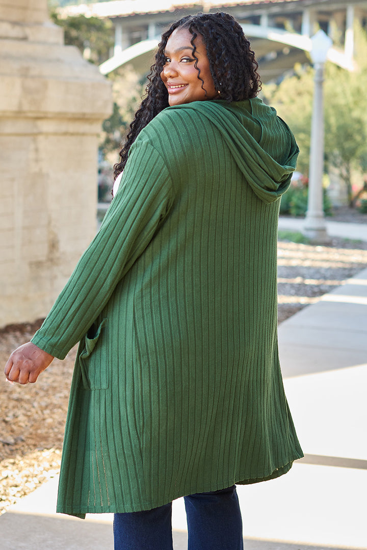 A person with long curly hair smiles while standing outdoors wearing the Basic Bae Full Size Hooded Sweater Cardigan in a lush green ribbed design, complete with subtle pockets. The backdrop of buildings and pathways hints at an urban park environment, ideal for the cardigan's opaque stretch fabric that ensures unrestricted movement.