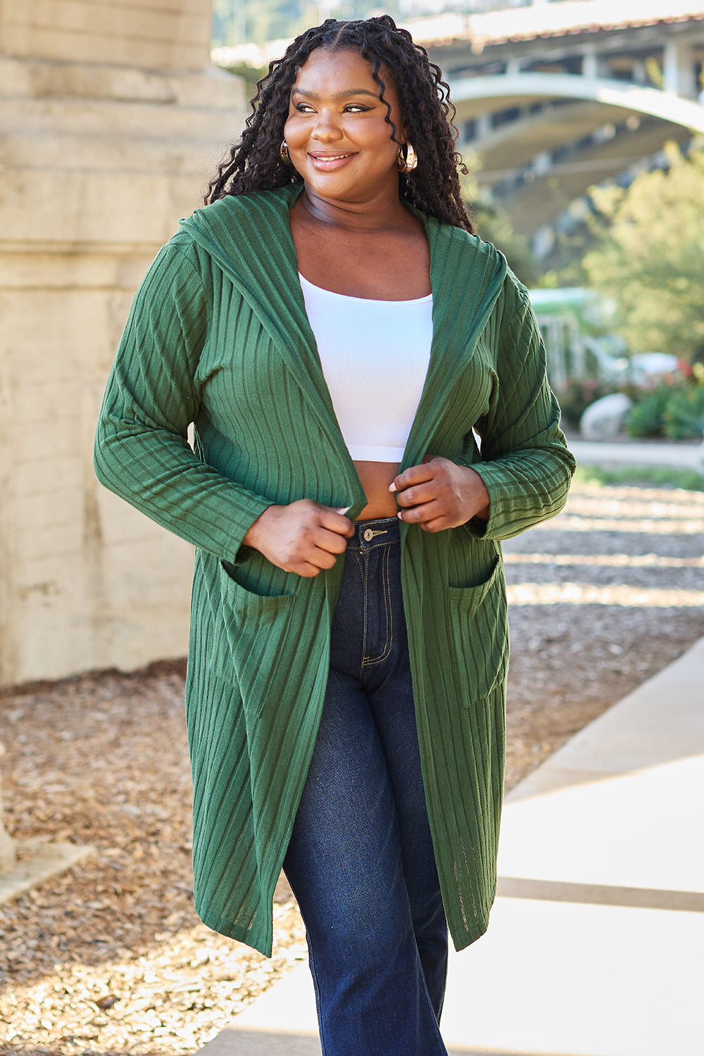 A woman beams outdoors, draped in the Basic Bae Full Size Hooded Sweater Cardigan over a white top and dark jeans. The sunlit setting with trees and a bridge in the distance accentuates her effortlessly chic style, perfected by the cardigan's pocketed sheer details.