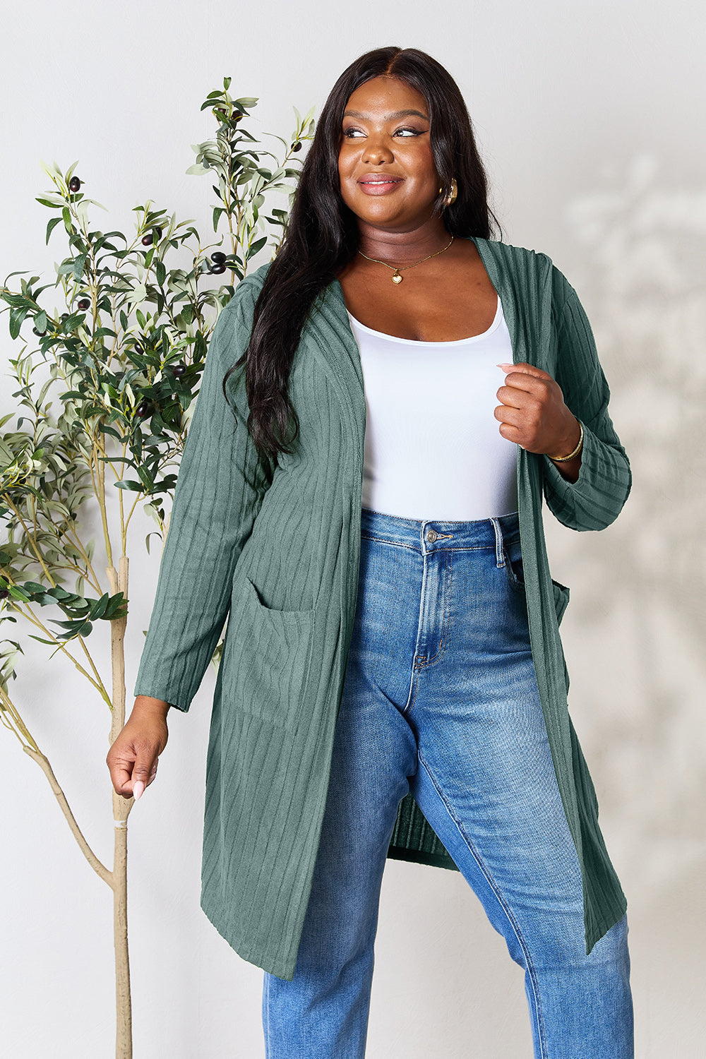 A woman smiling, wearing a Basic Bae Full Size Hooded Sweater Cardigan in green over a white shirt and blue jeans. She has long dark hair and stands in front of a light background with a green plant to her left. Her cardigan features an opaque stretch material, perfect for style and comfort.