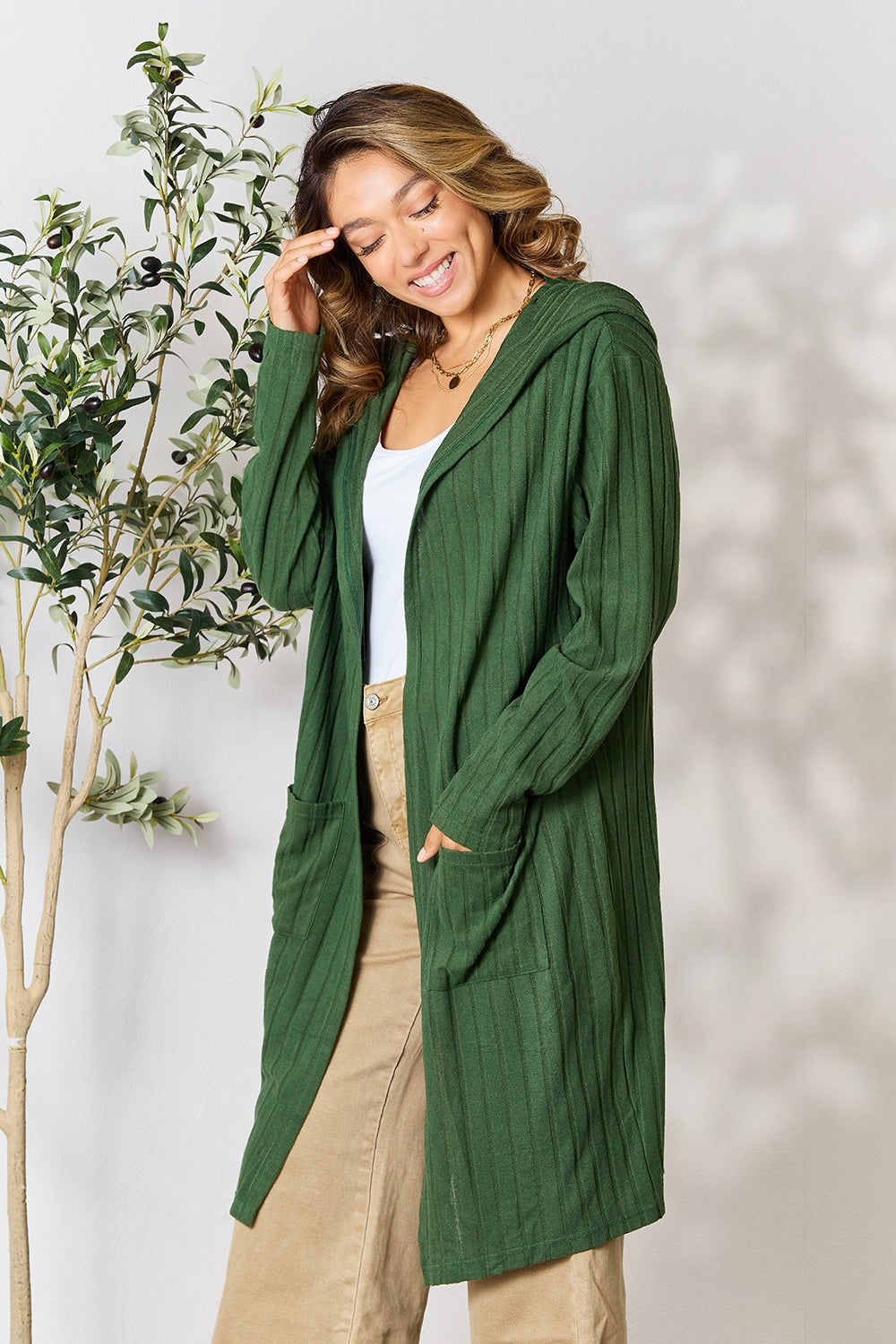 A person smiling and touching their hair, dressed in a green Basic Bae Full Size Hooded Sweater Cardigan, white top, and beige pants, stands next to a tall plant.