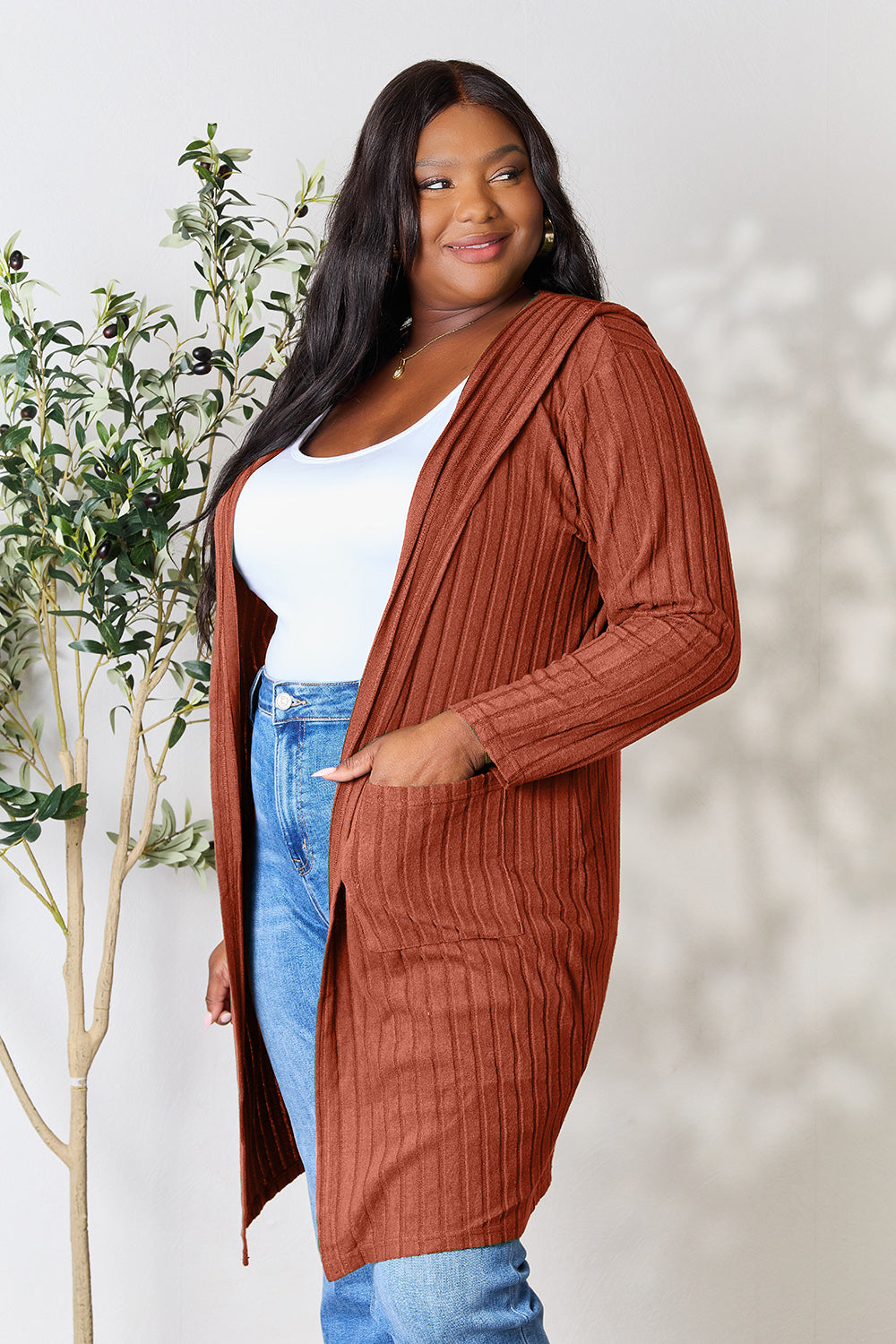 A person is smiling while wearing the Basic Bae Full Size Hooded Sweater Cardigan, a long, rust-colored layer over a white top and jeans. Standing in front of a light-colored wall with a small potted tree on the side, they enjoy the easy-care comfort of this opaque stretch fit cardigan—simply machine wash cold for convenience.