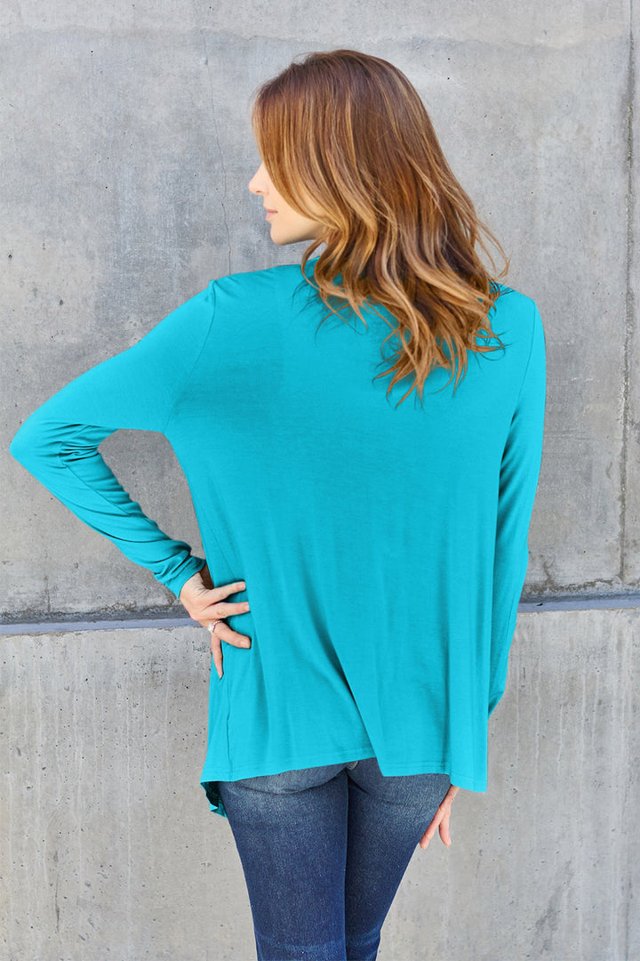 A person with long hair stands with their back to the camera, wearing a Basic Bae Full Size Open Front Long Sleeve Cardigan in teal, crafted from slightly stretchy rayon spandex, paired with jeans, against a concrete wall. Their left hand rests on their hip.