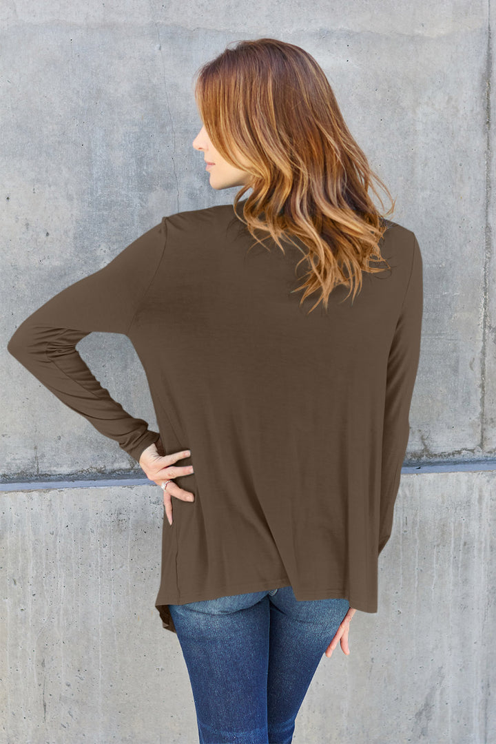 A woman with long, wavy hair faces away from the camera, wearing blue jeans and a Basic Bae Full Size Open Front Long Sleeve Cardigan in brown. She stands in front of a textured concrete wall with her hands on her hips, embodying a basic style enriched by the comfort of rayon spandex.