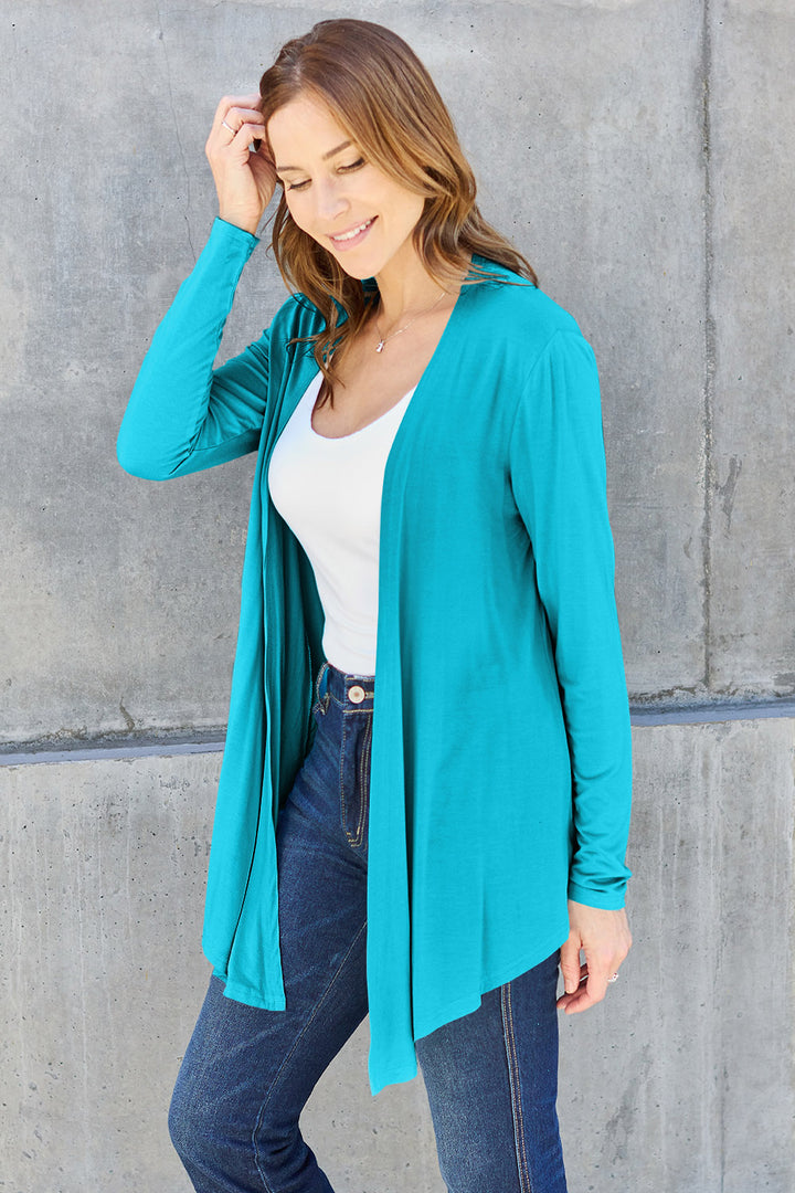 A woman in a white top, the Basic Bae Full Size Open Front Long Sleeve Cardigan in teal, and blue jeans crafted from slightly stretchy rayon spandex smiles while standing against a gray concrete wall. She holds a strand of her long brown hair with one hand, perfectly embodying a basic style.
