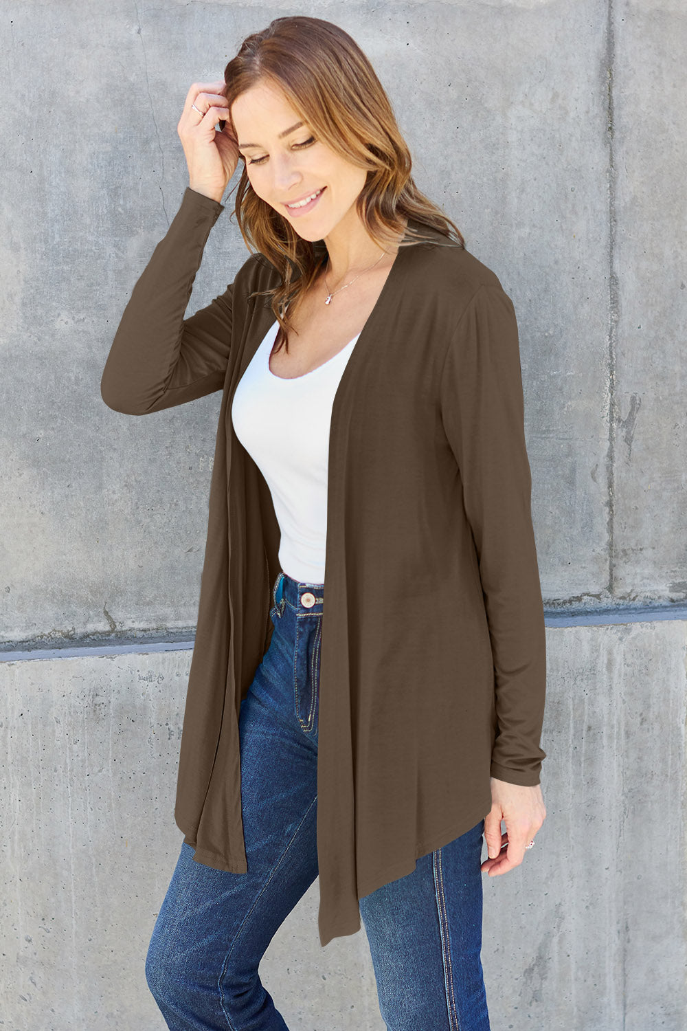 A person with long hair is smiling while posing against a concrete wall. They are wearing the Basic Bae Full Size Open Front Long Sleeve Cardigan, which is slightly stretchy and brown, made of rayon spandex, over a white top and blue jeans, with one hand touching their hair.