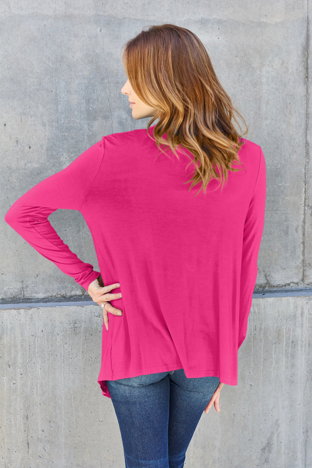 A person with long hair, wearing the Basic Bae Full Size Open Front Long Sleeve Cardigan in bright pink, made from slightly stretchy rayon spandex over jeans, faces a concrete wall with their back turned, and has one hand on their hip.
