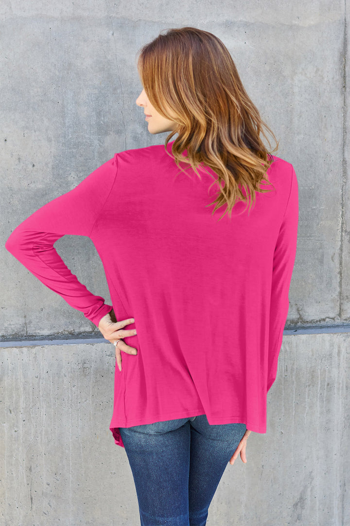 A person with long hair, wearing the Basic Bae Full Size Open Front Long Sleeve Cardigan in bright pink, made from slightly stretchy rayon spandex over jeans, faces a concrete wall with their back turned, and has one hand on their hip.