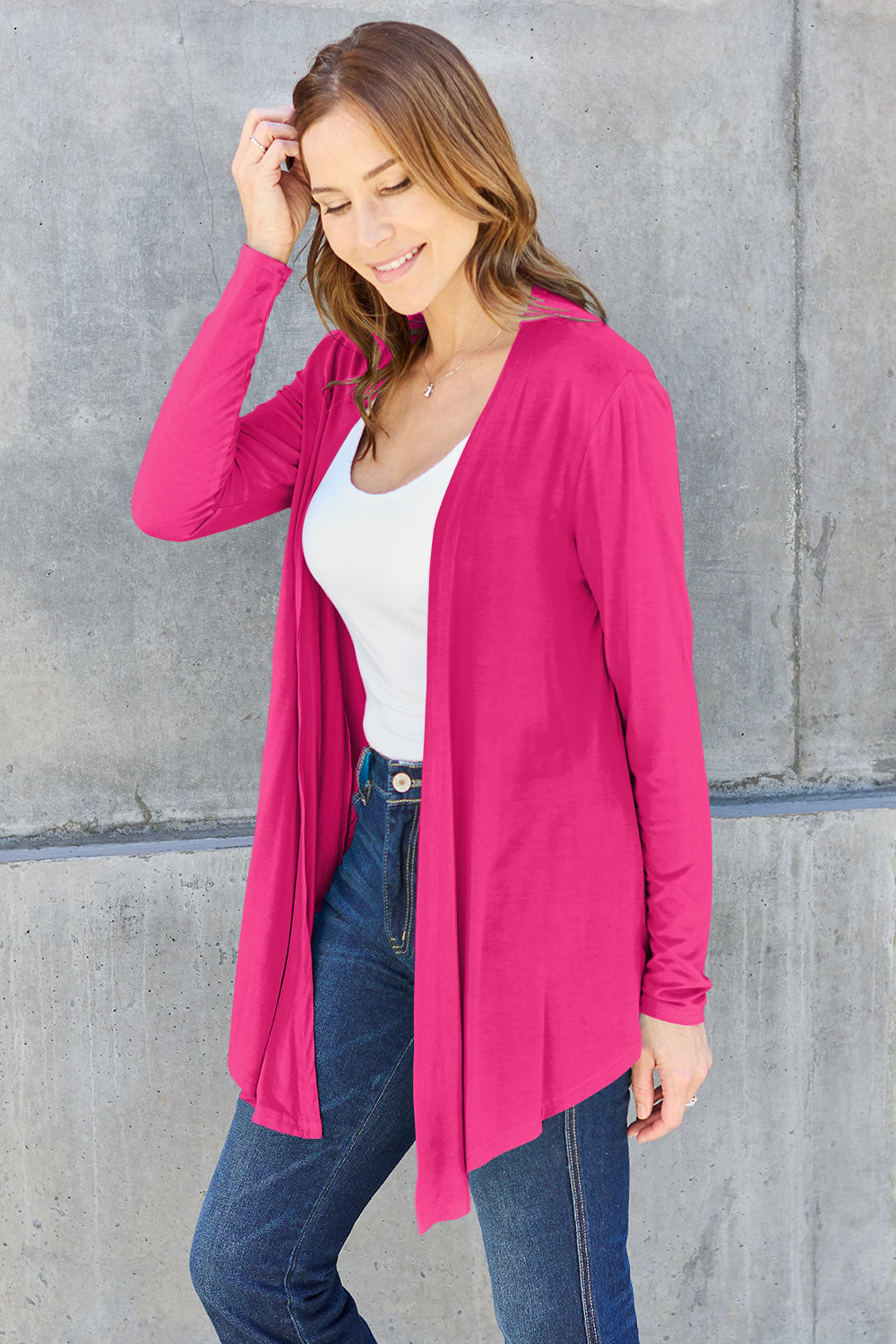 A woman stands against a concrete wall, embodying a basic style with her white top and blue jeans. The Basic Bae Full Size Open Front Long Sleeve Cardigan, crafted from slightly stretchy rayon spandex in a bright pink hue, adds flair. Her long brown hair cascades as she smiles, casually touching it with one hand.