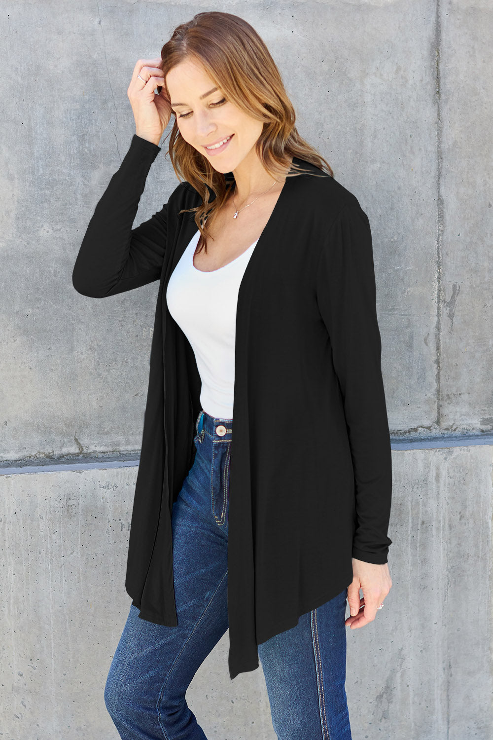 A woman with long hair stands in front of a concrete wall, wearing the Basic Bae Full Size Open Front Long Sleeve Cardigan in black, paired with a white top and blue jeans. Her basic style is effortlessly chic as she smiles while touching her slightly stretchy outfit with one hand, striking a relaxed pose.
