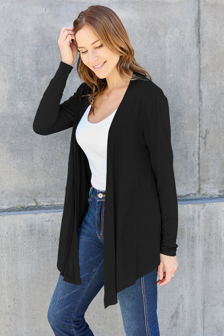 A woman with long hair stands in front of a concrete wall, wearing the Basic Bae Full Size Open Front Long Sleeve Cardigan in black, paired with a white top and blue jeans. Her basic style is effortlessly chic as she smiles while touching her slightly stretchy outfit with one hand, striking a relaxed pose.