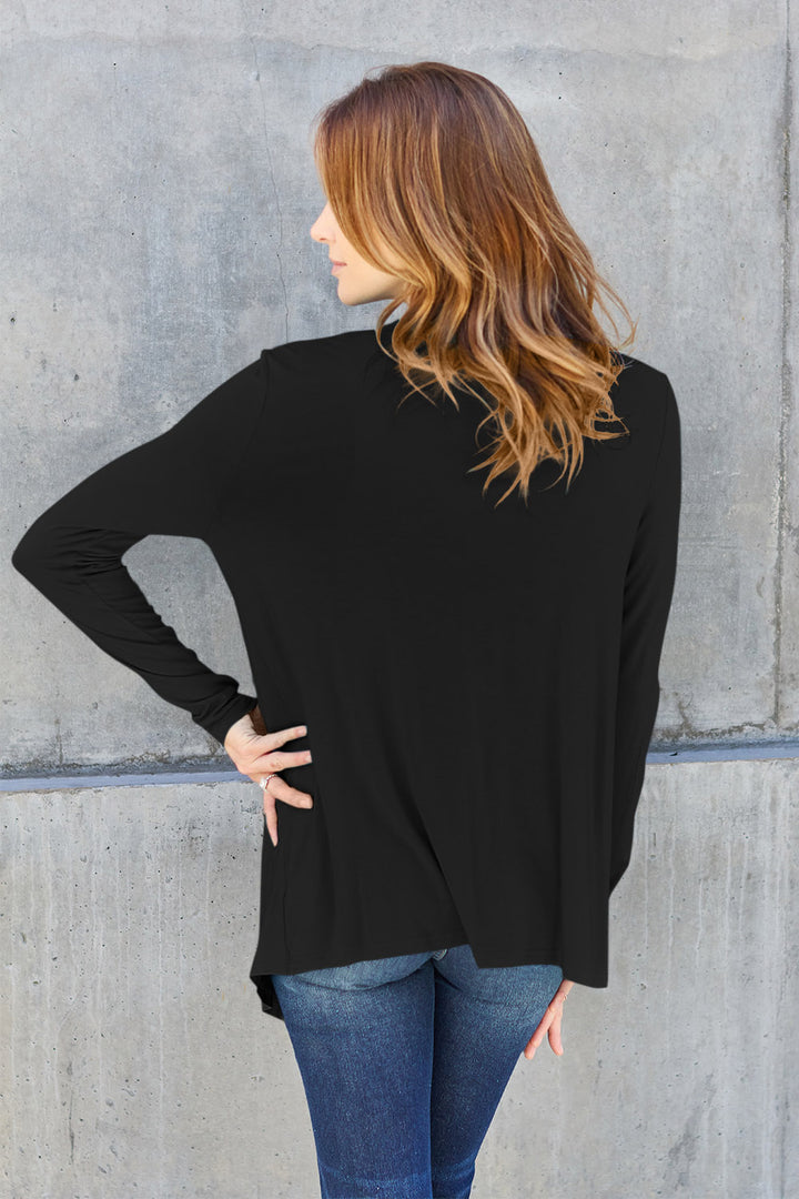A person with long, wavy hair stands with their back to the camera, wearing a Basic Bae Full Size Open Front Long Sleeve Cardigan in black and blue jeans, posing against a textured gray wall.