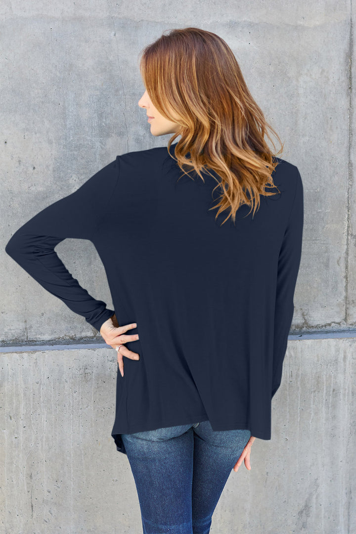 A woman with long, wavy hair stands with her back to the camera, wearing the Basic Bae Full Size Open Front Long Sleeve Cardigan in navy blue paired with jeans. Her right hand is on her hip, as she poses in front of a concrete wall.