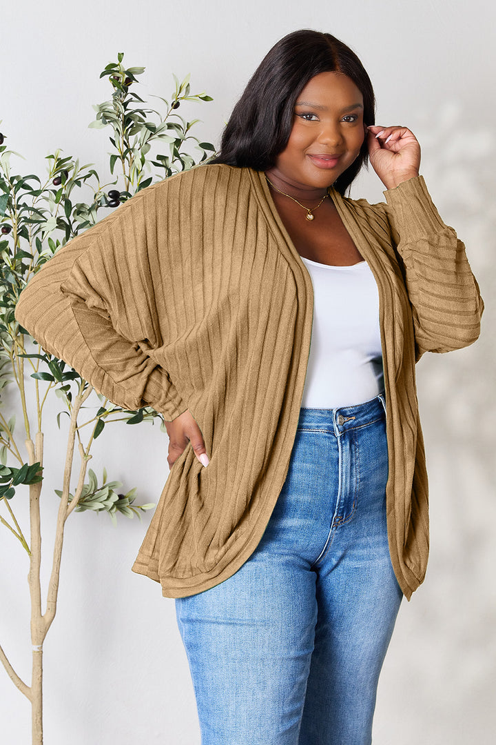 A person is smiling and posing in front of a light background, wearing the Basic Bae Full Size Ribbed Cocoon Cardigan in beige over a white top, paired with blue jeans. A small plant in the background complements the minimalist style of the setting.