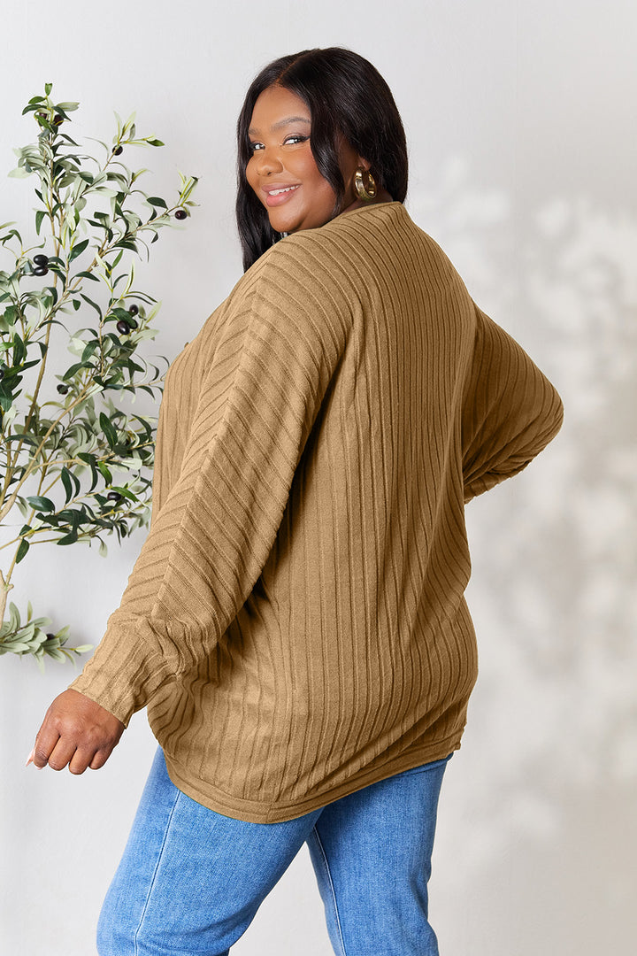 A woman with long dark hair is smiling and standing in a relaxed pose, showcasing her basic style. She is wearing a slightly stretchy, ribbed camel-colored Basic Bae Full Size Ribbed Cocoon Cardigan along with blue jeans. A leafy plant is partially visible in the background on the left side.