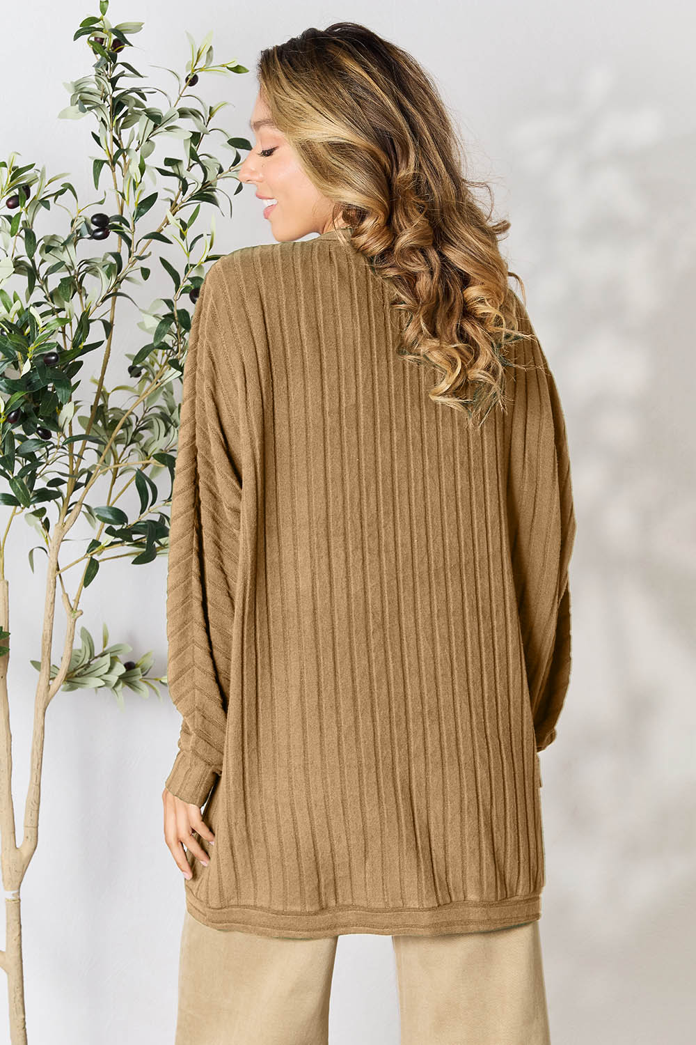 A person with long wavy hair is wearing the Basic Bae Full Size Ribbed Cocoon Cardigan in a camel color, paired with beige pants. Embracing a casual style, they stand next to a leafy plant, facing away from the camera, while the background is softly blurred.