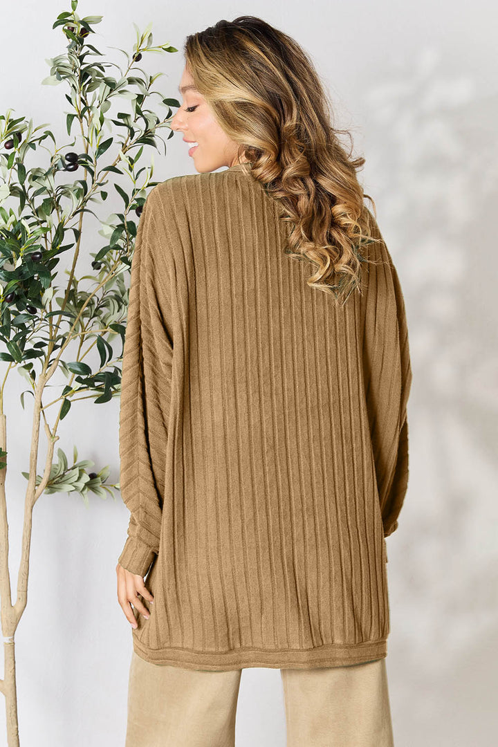 A person with long wavy hair is wearing the Basic Bae Full Size Ribbed Cocoon Cardigan in a camel color, paired with beige pants. Embracing a casual style, they stand next to a leafy plant, facing away from the camera, while the background is softly blurred.