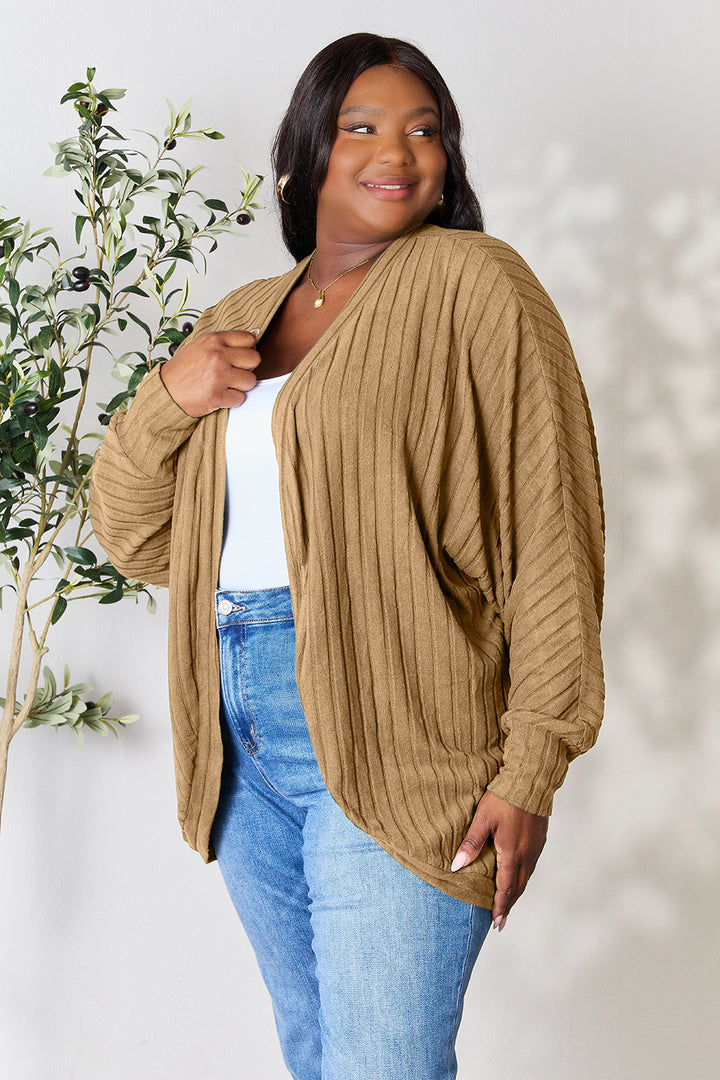 A woman stands smiling in her basic style, wearing the Basic Bae Full Size Ribbed Cocoon Cardigan in camel over a white top and blue jeans. She is posed against a light-colored wall with a leafy plant by her side, exuding effortless charm.
