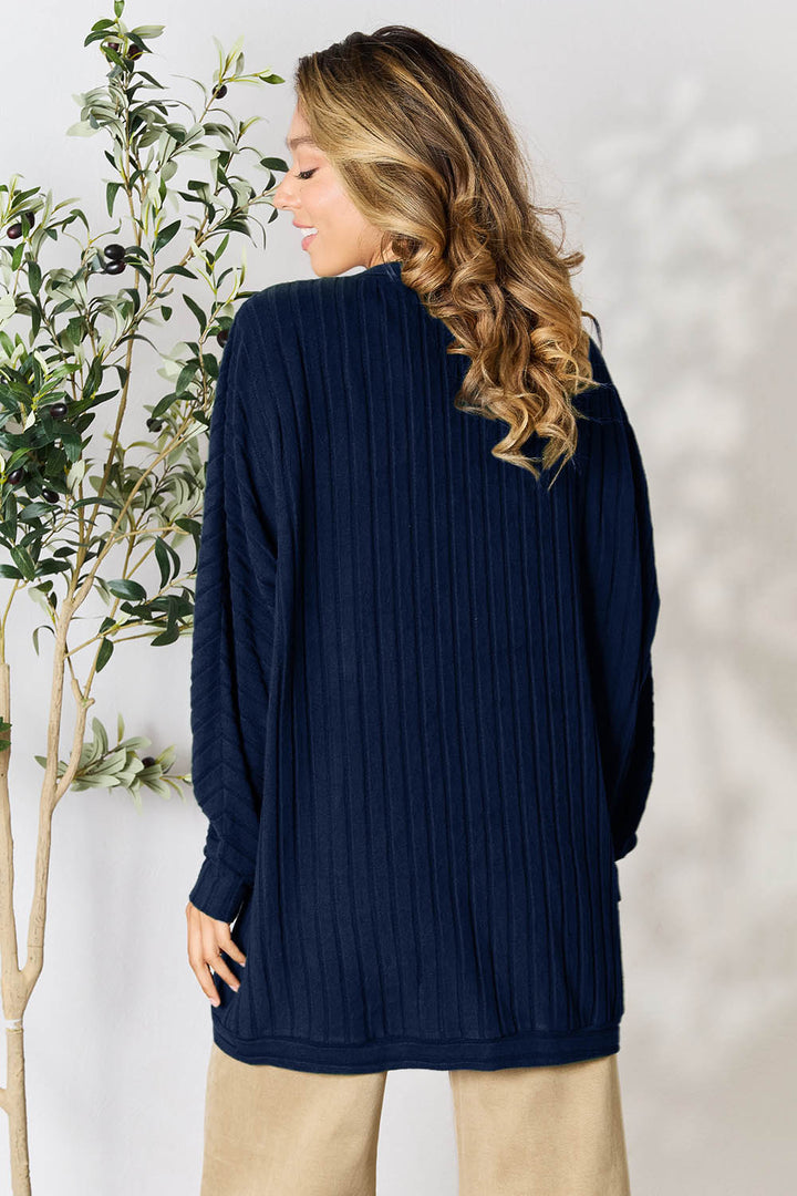 A person with wavy, light brown hair stands facing away, wearing the Basic Bae Full Size Ribbed Cocoon Cardigan in dark blue and slightly stretchy beige pants. They are next to a leafy plant in a neutral-toned room.