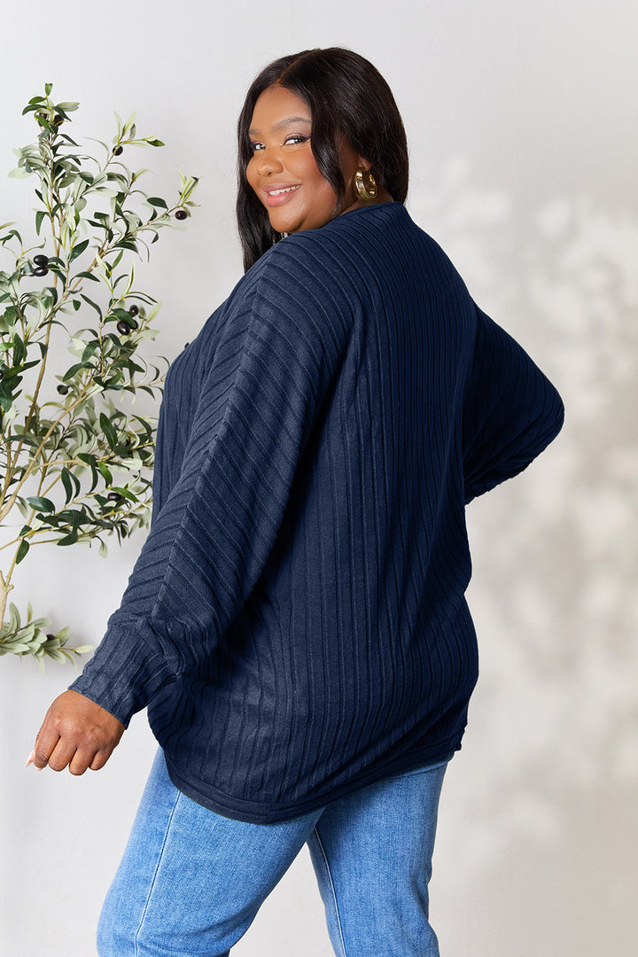 A smiling person with long dark hair, wearing a Basic Bae Full Size Ribbed Cocoon Cardigan made from slightly stretchy polyester and light blue jeans, stands in front of a light background with a plant to the side.
